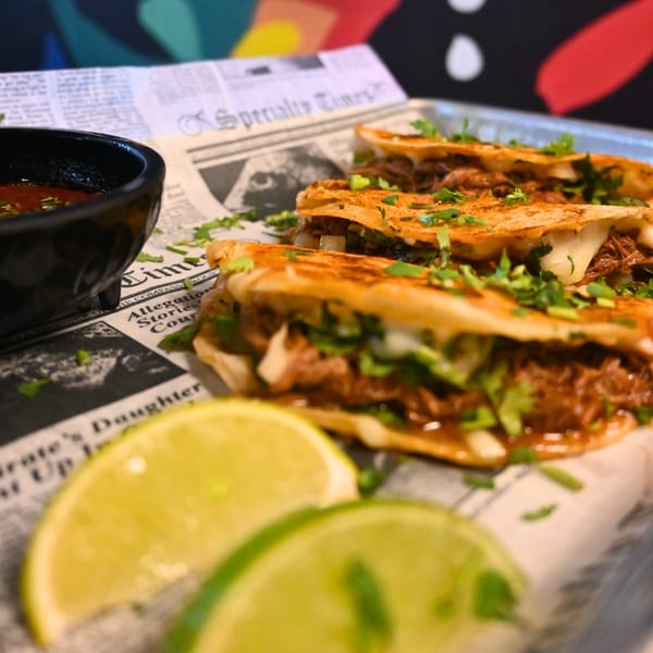 Three La Reyna Xpress birria tacos with cilantro, a side of consommé, and lime wedges.