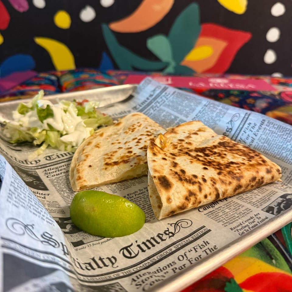 Two quesadillas with a side of lettuce, sour cream, and lime on newspaper-lined tray.