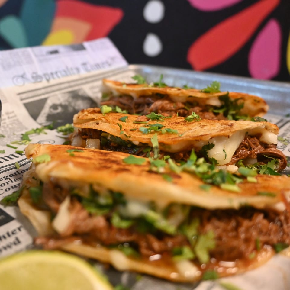 Three La Reyna Xpress birria quesadillas with melted cheese and cilantro on newspaper-lined tray.