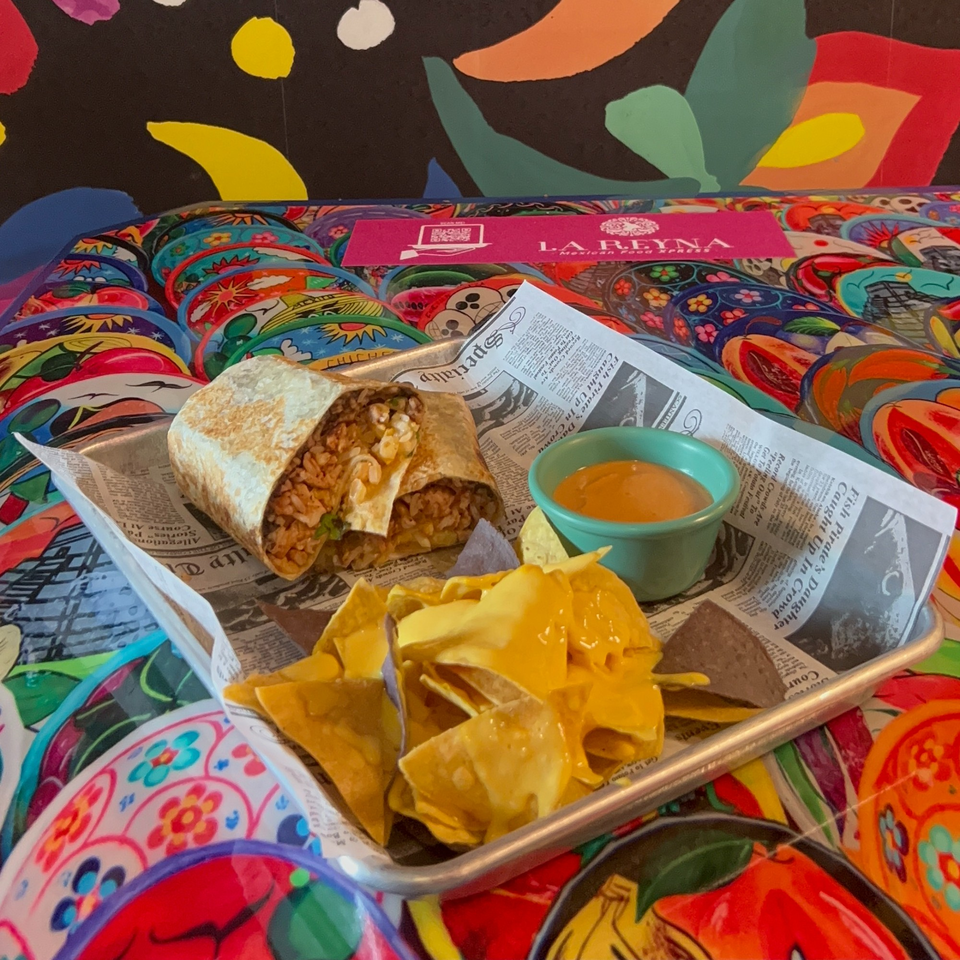 La Reyna Xpress burrito, nachos with cheese, and dipping sauce on a colorful table.