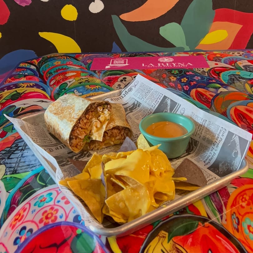 La Reyna Xpress burrito, nachos with cheese, and dipping sauce on a colorful table.