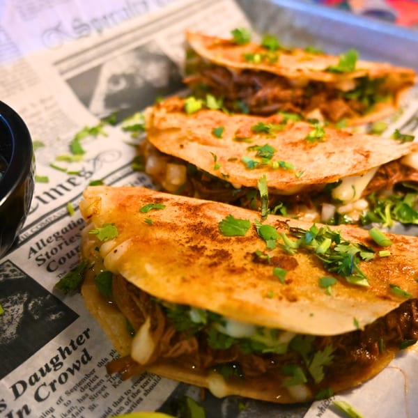 Three La Reyna Xpress birria tacos with cilantro and onions on newspaper-lined tray.