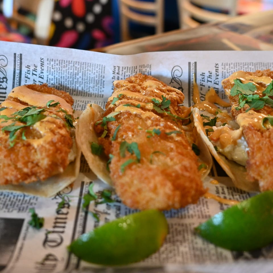Three crispy fish tacos with sauce and cilantro, served on newspaper-print paper with lime wedges.