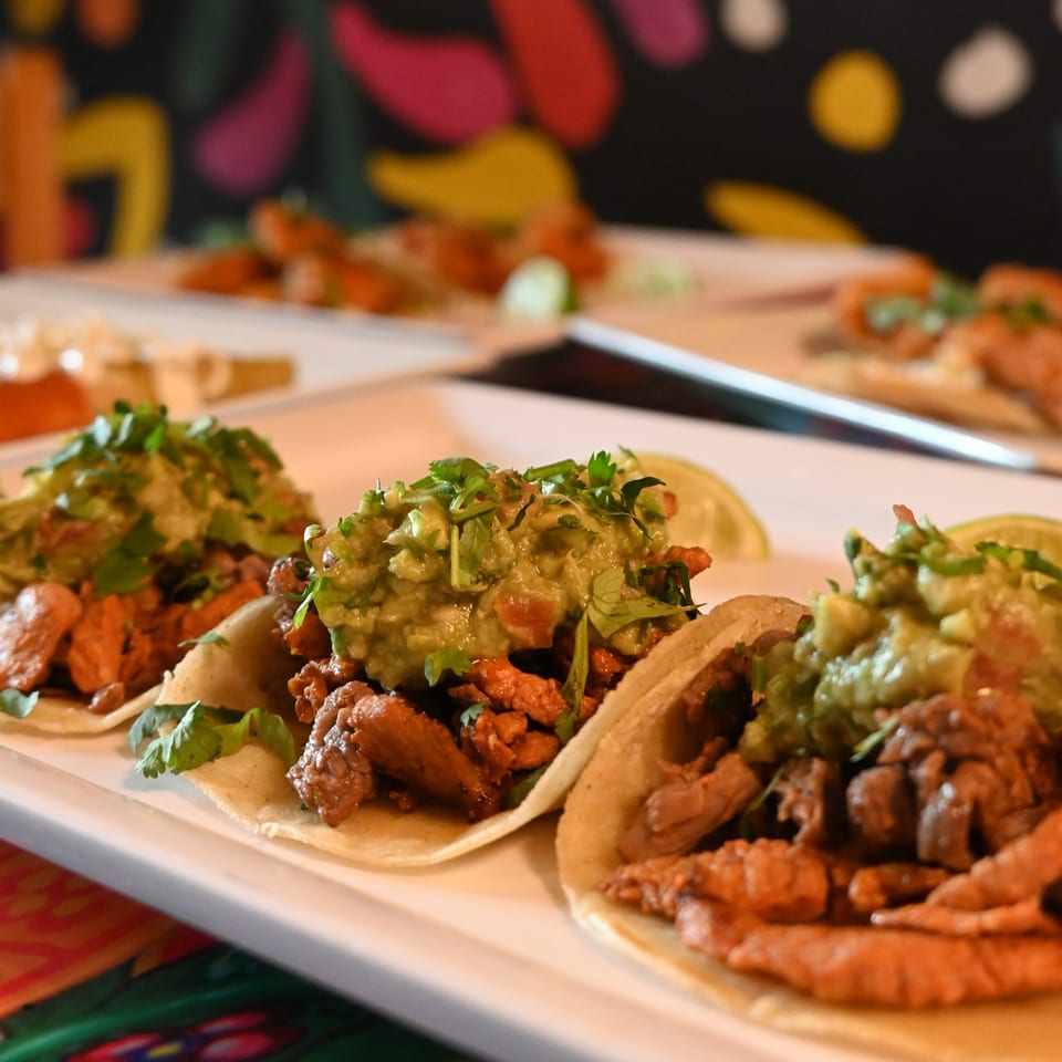 Three La Reyna Xpress tacos with meat, guacamole, and cilantro on a white plate.