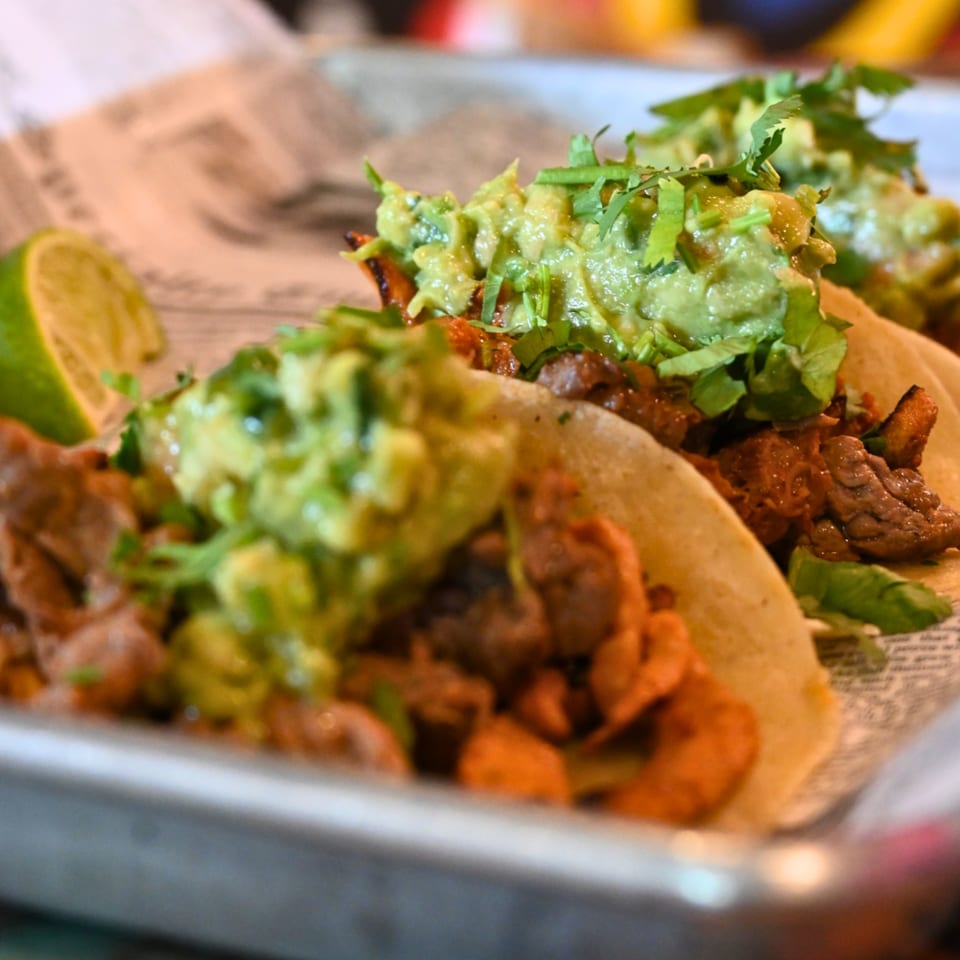 Three La Reyna Xpress tacos with meat, guacamole, and cilantro, served with a lime wedge.