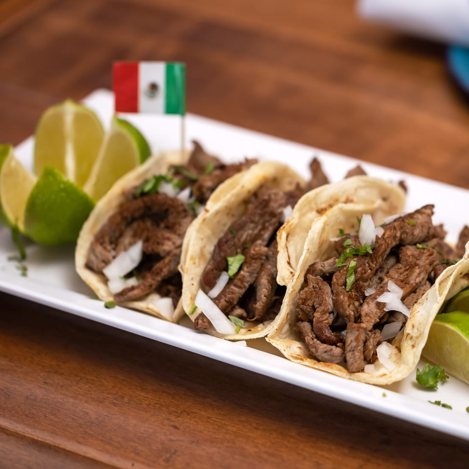 Three La Reyna Xpress carne asada tacos with onions, cilantro, lime, and a Mexican flag.